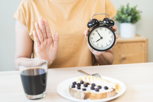 Asian young woman raising hand in stop gesture refusing healthy breakfast food while holding alarm clock, intermittent fasting weight loss regimen and time restricted eating nutrition concept.