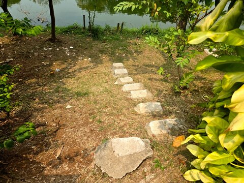 ​Concrete stepping stones path through a grassy garden park down towards a calm lake, surrounded by green trees and tropical foliage