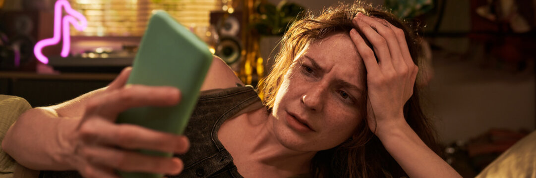 Stressed Caucasian young adult woman looking at smartphone screen with worried expression. Image represents cyberbullying, bad news, or digital fatigue for mental health campaigns