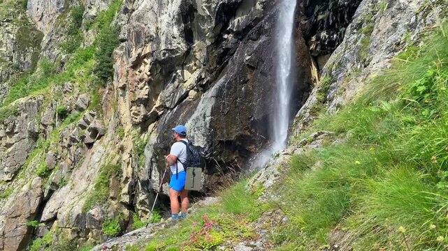 Male hiker observing waterfall in wide view on rocky slope near Ushba, Svaneti, Georgia, travel scene for trekking promotion, adventure tourism, route marketing, and mountain destination campaigns