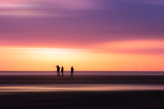 Aerial view of three silhouettes standing on a beach at sunset with motion blur effects on the sand and sky in Sligo, County Sligo, Ireland.