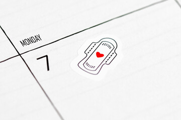 Close up of calendar with sticker showing sanitary pad and red heart. Reminder for tracking menstrual cycle and period start © Firn