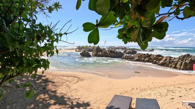 Wide shot of a beautiful Sri Lankan beach surrounded by lush greenery, featuring large breakwaters and old sunbeds on the sandy shore during daytime.