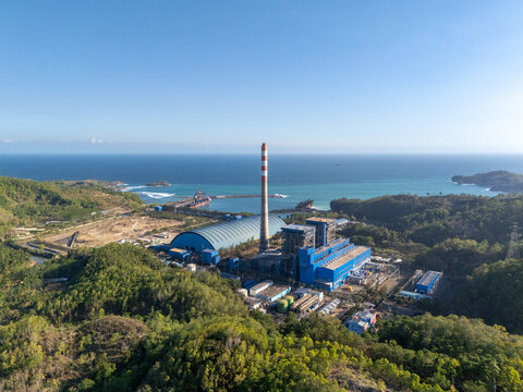 Aerial view of the power plant with its tall chimney and blue industrial buildings along the lush green coastline overlooking the sea in Pacitan, East Java, Indonesia.