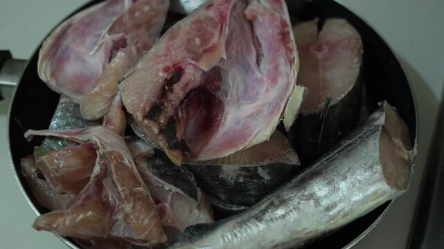 Close-up shot of raw fish pieces in a frying pan, ready for cooking. The fresh seafood highlights traditional coastal cuisine preparation, with natural lighting and kitchen setting.
