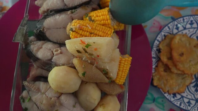 Close-up shot of authentic Colombian sancocho featuring fish, yuca, corn, and potatoes in a glass dish, beside crispy golden patacones on a decorative plate. Natural daylight enhances colors.