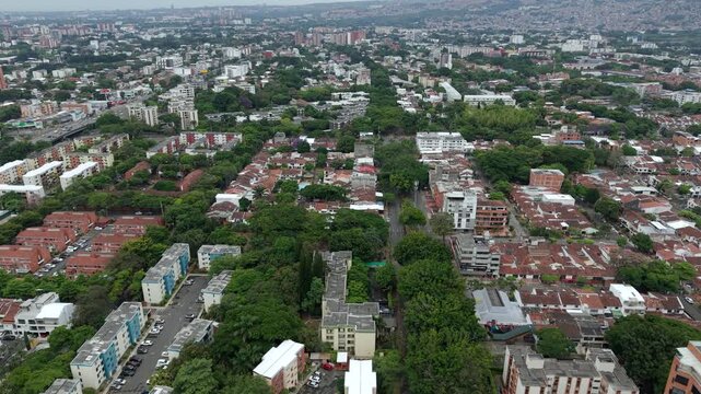 Drone aerial footage reveals the vibrant Calle Novena area in Cali, Colombia, featuring residential neighborhoods, abundant trees, diverse buildings, and cityscape under cloudy daylight.