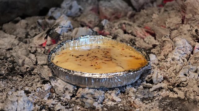 Traditional Argentine provoleta cheese cooks in a small pan over glowing charcoal during an outdoor barbecue.