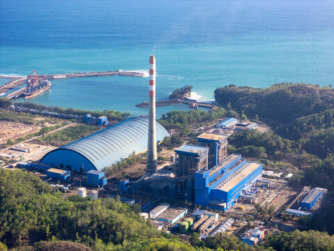 Aerial view of the Pacitan Power Plant with its tall chimney, blue storage dome, and industrial buildings along the coast with a cargo ship docked in Pacitan, East Java, Indonesia.