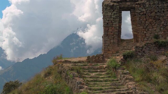 Static wide shot of an ancient stone doorway ruin and steps on a cliff edge with dramatic clouds and snow capped mountains in Cusco, Peru.