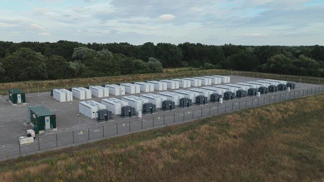Multiple industrial battery units are arranged in rows within a fenced outdoor site as natural daylight fades during the early evening hours in a quiet countryside landscape