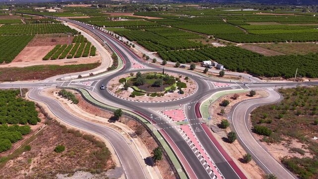 Large roundabout intersection surrounded by agricultural fields and orchards viewed from above, featuring red cycle lanes, road markings and sparse traffic in rural Spain