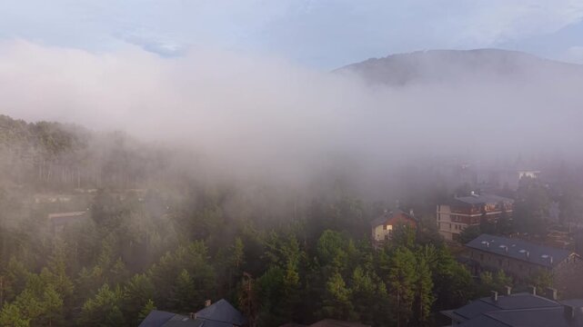 Cloud inversion sweeps over La Molina mountain village in Spain, with low fog rolling through pine forests and alpine buildings beneath a clearing blue sky