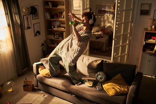 Young adult Caucasian man wearing headphones dancing on sofa and drinking beer in messy room. Image represents youth lifestyle and is suitable for beverage advertising, high angle shot
