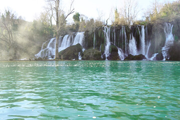Wide view of turquoise lake with cascades at Kravica Waterfall. Depicts Bosnia and Herzegovina's iconic karst landmark at low season, travertine tufa formation, and raw power of Trebižat River. © tanyatorgonskaya