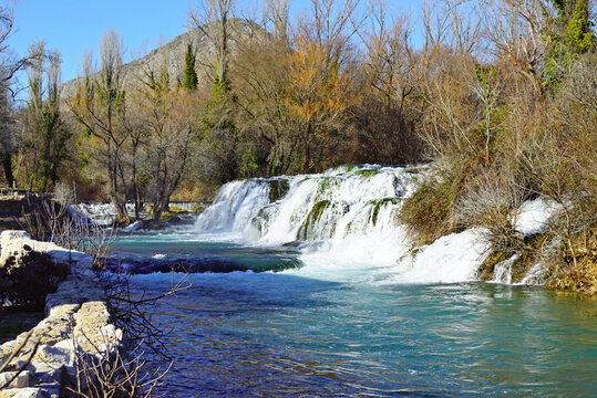 Wide cascade of Kocusa Waterfall flowing into clear turquoise river surrounded by trees. Captures travertine barrier hydrology, dramatic Balkan natural spectacle, and water resources preservation.