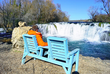 Person relaxing on wooden bench overlooking wide roaring cascades at Kocusa Waterfall. Symbolizes contemplative nature tourism, slow travel through Herzegovinian karst sites, and off-season solitude. © tanyatorgonskaya