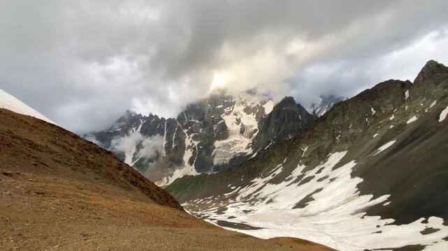 Clouds enveloping Mount Ushba in wide view above lingering snowfields in Svaneti, Georgia, mountain scene for alpine climate media, route assessment, seasonal snow coverage, and expedition planning.