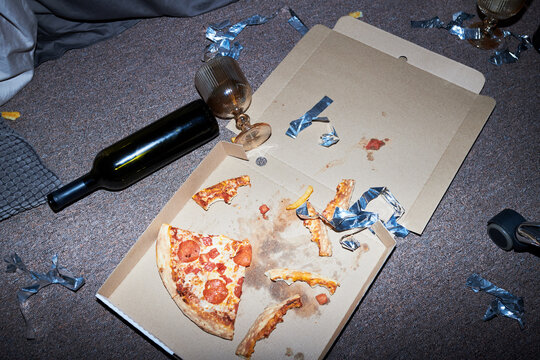 Messy floor after party showing leftover pizza in cardboard box, empty wine bottle, and glasses. High angle view perfect for youth lifestyle, student housing, or cleanup service marketing