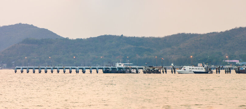 Long jetty and moored boats in evening light at Dongtan Gulf, Sattahip. Panoramic view of concrete pier with docked boats illuminated by streetlights against hills in Chon Buri, Thailand during dusk.