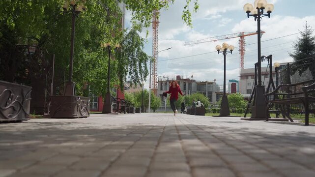 Woman in red coat walks along paved pathway lined with ornate street lamps and greenery holding object. background features buildings under construction or renovation alongside existing