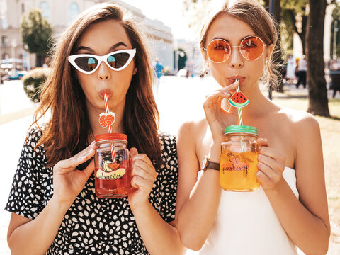 Two young beautiful smiling hipster in trendy summer clothes,dress.Sexy carefree women posing in the street.Positive models having fun in sunglasses.Drinking fresh cocktail smoozy drink