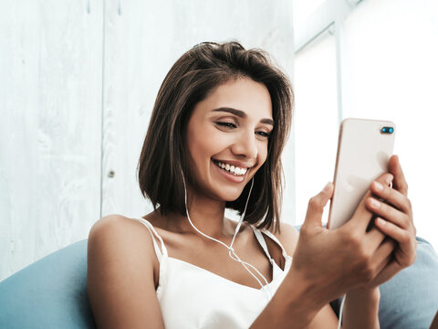 Portrait of beautiful smiling woman dressed in white pajamas and listening to music with headphones. Carefree model sitting on soft bag chair. Sexy female enjoying her morning at balcony