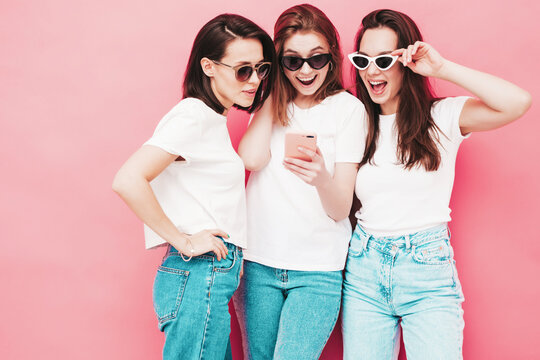 Three young beautiful smiling hipster female in trendy summer  t-shirt and jeans clothes. Sexy carefree women posing near pink wall in studio.Positive models looking at smartphone screen. Using apps