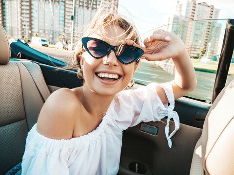 Portrait of young beautiful and smiling hipster in convertible car. Sexy carefree woman driving cabriolet. Positive model riding and having fun in sunglasses at sunset