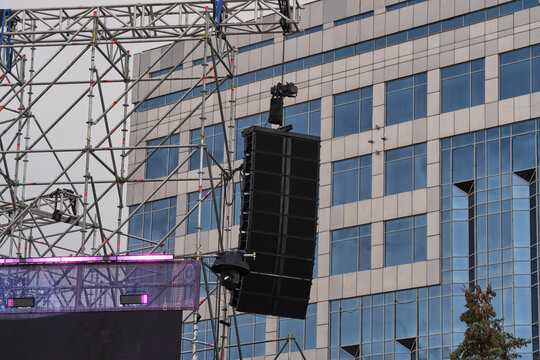 Outdoor concert stage rigging with hanging speakers and lights set up against modern glass building.
