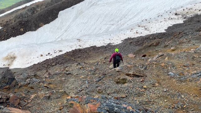 Hiker descending in rear view on a rocky mountain slope near summer snowfields in Svaneti, Georgia, for trekking safety, alpine route planning, endurance travel, and expedition training use