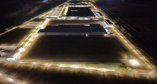 Aerial view of the illuminated ZF Friedrichshafen AG industrial plant at night, featuring modern factory buildings, extensive parking areas, and surrounding fields in Pancevo, Serbia.