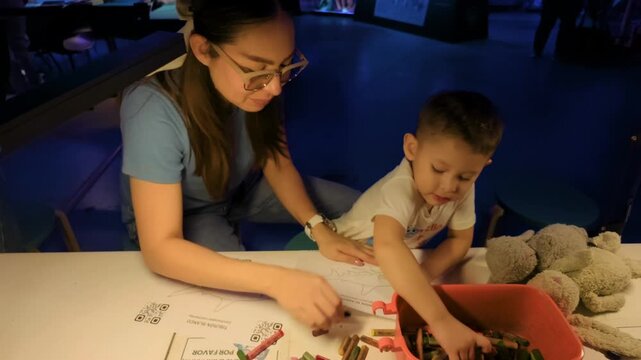 A young Latina mother and her 2-year-old baby use crayons to color shark-themed pages in an interactive exhibit, illuminated by blue LED lights, creating an underwater ambiance.