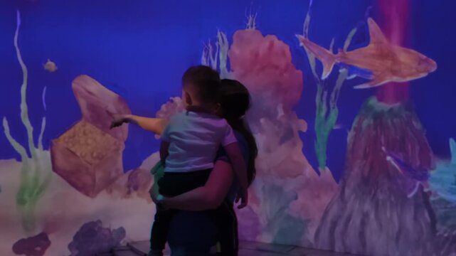 A mother holds her baby as they observe interactive digital projections of sharks and other marine life on a wall, an engaging experience at a modern aquarium exhibit.