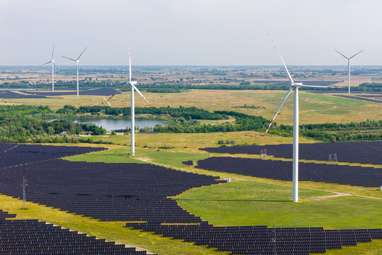 Aerial view of a large solar farm with rows of photovoltaic panels and wind turbines set in a green landscape with a small lake in Poland.
