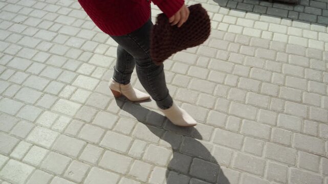 Close up of woman walking on light gray brick pavement wearing red sweater dark jeans and white ankle boots while holding brown knitted bag. Foot movement is captured from various angles