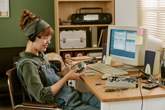 Young adult Caucasian woman wearing headphones and holding portable CD player in retro office. Image represents vintage technology and is suitable for nostalgic marketing campaigns