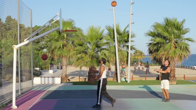 Two men playing basketball and shooting hoops on an outdoor court by the sea