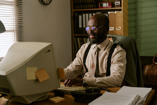 Middle aged African American man working on retro computer in vintage office, typing on keyboard and smiling. Image represents corporate history and nostalgia for marketing campaigns