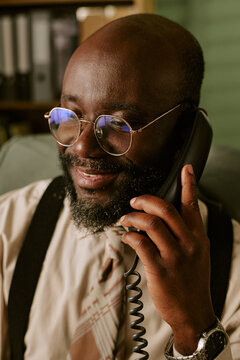 Vertical image of middle aged African American man smiling while talking on retro landline telephone in office, representing vintage corporate communication and customer service