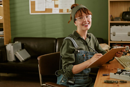 Portrait of smiling young adult Caucasian woman technician writing on clipboard in retro office. Image represents vintage engineering and is suitable for technical education marketing