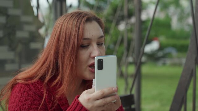 Close up of woman applying lip gloss while looking at her smartphone in outdoor park setting. Woman holds phone and applicator with greenery visible in soft focus background