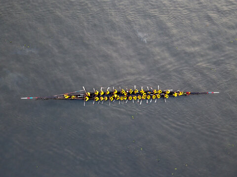 Aerial view of a traditional boat race featuring a long, narrow racing boat with many rowers in yellow shirts paddling on the river in Gopalganj, Dhaka Division, Bangladesh.