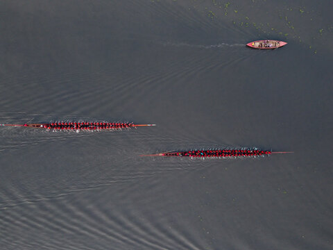 Aerial view of a traditional boat race with long, narrow boats filled with rowers in red shirts on a dark river in Gopalganj, Dhaka Division, Bangladesh.