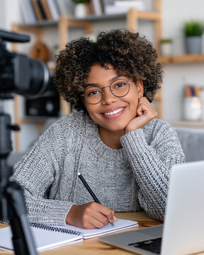 Young woman smiles warmly while writing notes. She wears glasses and a cozy sweater. Laptop and notebook sit beside her. Camera records her in front of a wooden shelf. She appears focused and cheerful