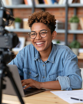 Warm smile radiates from joyful young woman. Curly hair and glasses frame her cheerful expression. She types on laptop beside professional camera setup. Wooden desk holds papers, pen
