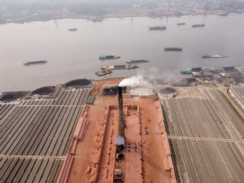 Aerial view of a brick kiln with a smoking chimney situated by the river, surrounded by rows of drying bricks and cargo boats navigating the water in Dhaka, Bangladesh.