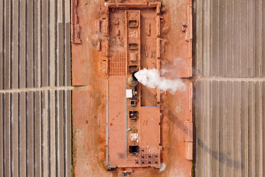Aerial view of a brick kiln with smoke rising from a central chimney, surrounded by rows of drying bricks in Dhaka, Bangladesh.