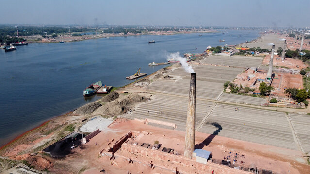 Aerial view of a brick kiln with a smoking chimney and rows of drying bricks along the riverbank, Dhaka, Bangladesh.