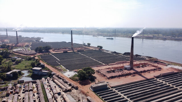 Aerial view of the brick kiln located by the river in Dhaka, Bangladesh, featuring tall chimneys emitting smoke, rows of drying bricks, and nearby industrial structures.